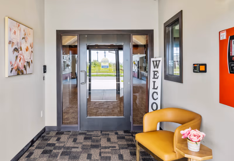 Interior entrance lobby with glass doors, a vertical 'WELCOME' sign, and a yellow chair beside a small table with flowers.