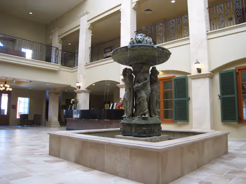 Spacious interior lobby area with a large decorative fountain featuring sculpted figures in the center. The lobby has beige tiled floors, tall columns, and a second-floor balcony with wrought iron railings. There are green shuttered windows on the walls and a reception desk in the background.