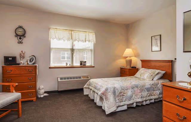 A cozy bedroom with a single bed covered in a patchwork quilt. The room has wooden furniture including a dresser, nightstand with a lamp, and a chair. A window with lace curtains lets in natural light, and a wall clock and framed pictures decorate the walls.