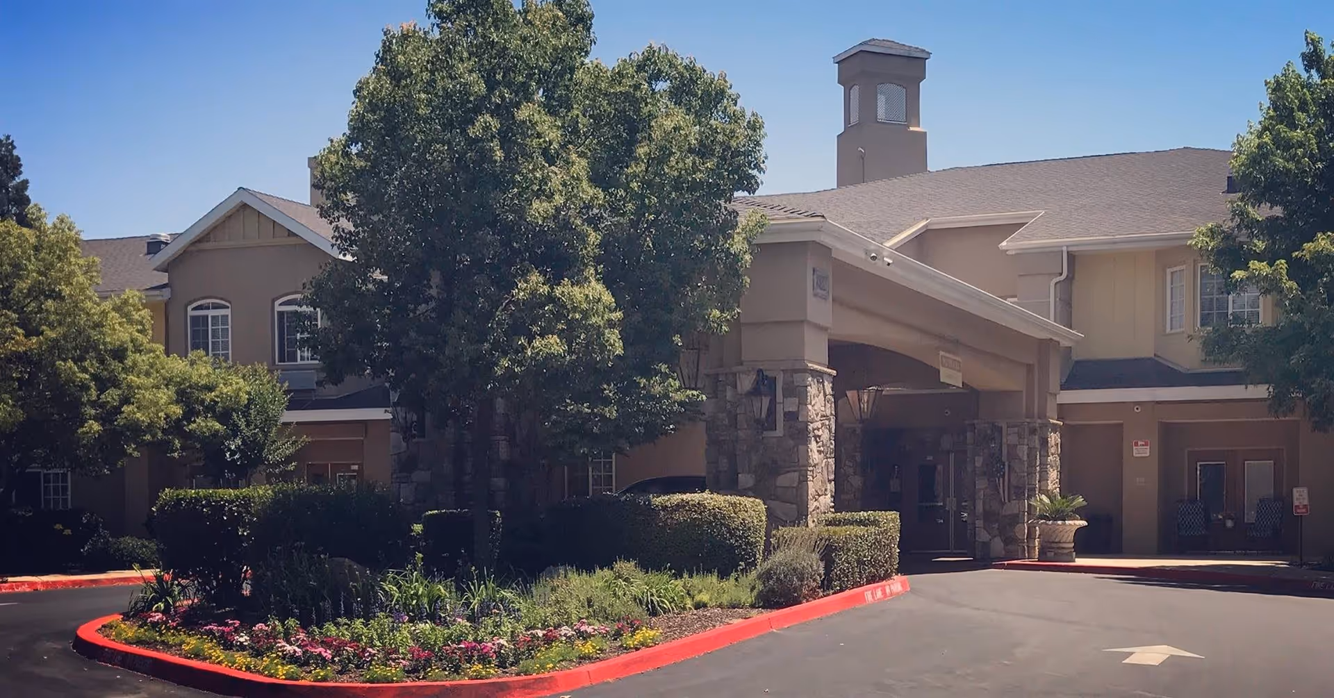 Front entrance of a senior living building with a covered porte-cochère, stone columns, trees and landscaped flower beds.