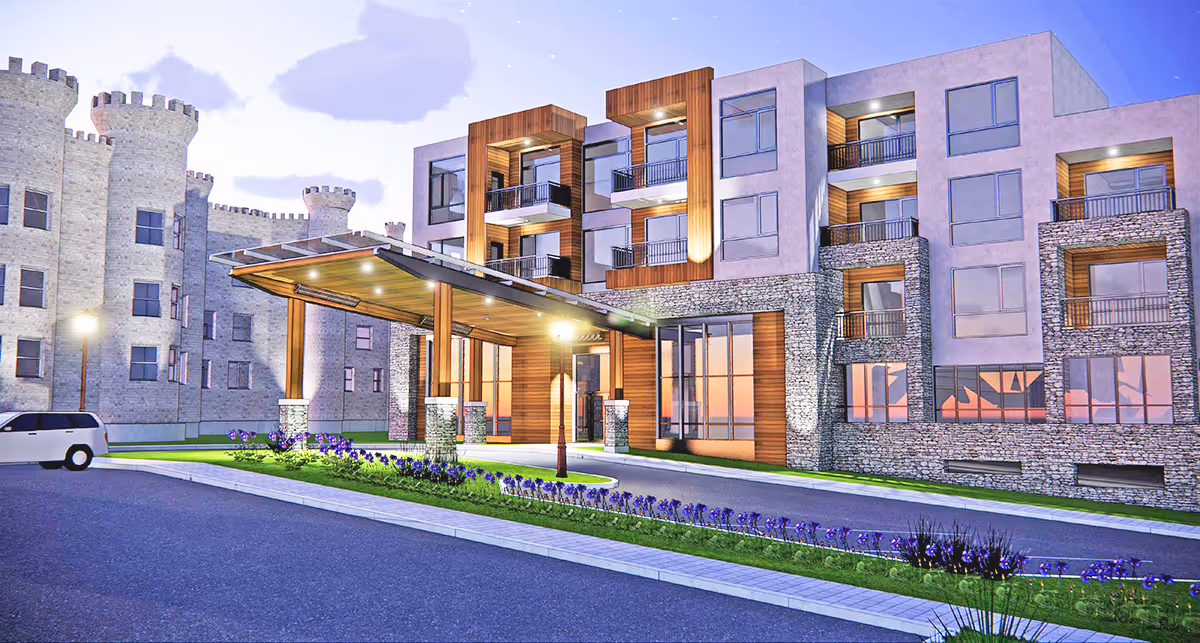 Exterior view of a modern senior living facility building with stone and wood accents, multiple balconies, and a covered entrance. The building is illuminated with outdoor lights during dusk, and there is a driveway with a white vehicle parked on the left side. Purple flowers line the median strip in front of the entrance.