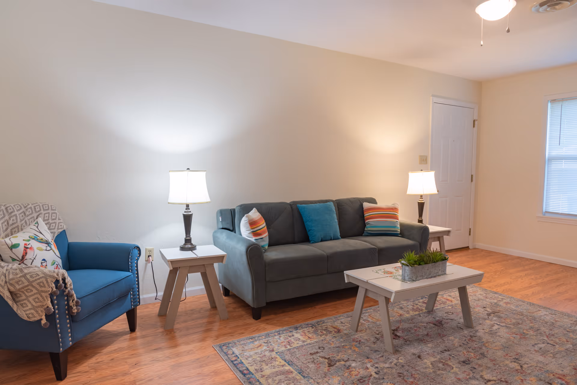 A cozy living room with a gray sofa adorned with colorful pillows, a blue armchair with a patterned throw blanket, two side tables each with a lamp, a white coffee table with a small plant, a patterned area rug, and a window with blinds letting in natural light.