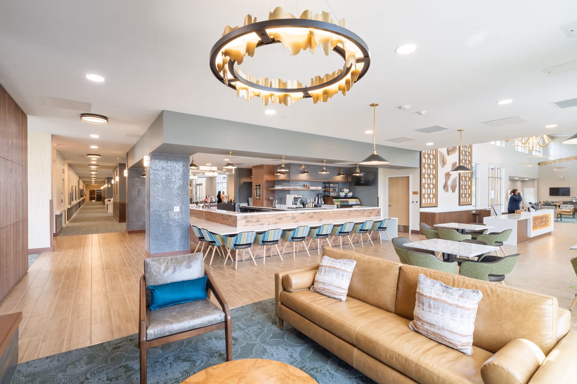 A spacious lobby area featuring a modern design with a large chandelier, comfortable seating, and a bar area in the background. The floor is tiled with wooden accents, and there are tables and chairs arranged for dining or socializing.