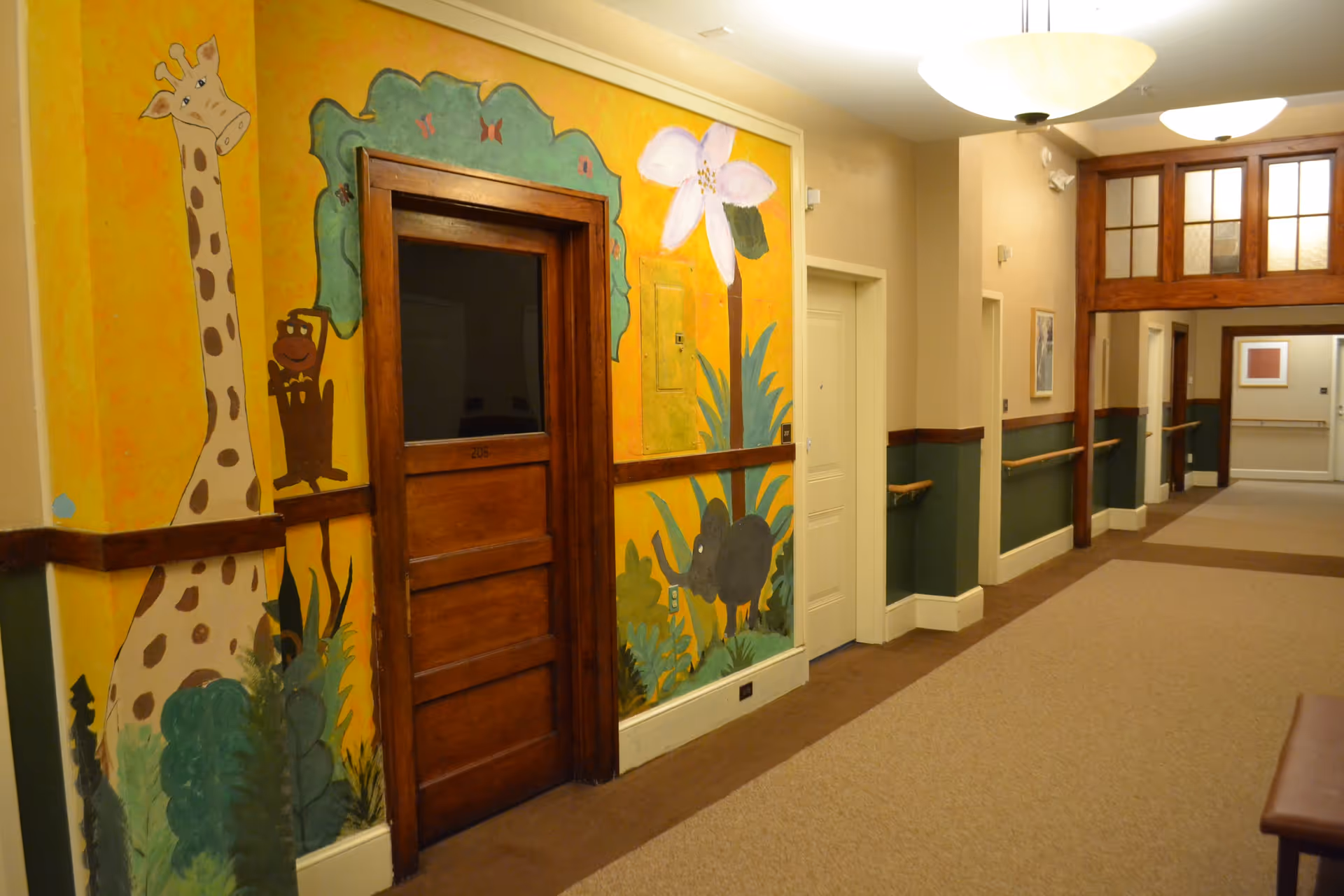 A hallway in a senior living facility with beige walls and carpeted floor. One wall features a colorful mural with a giraffe, a flower, and other plants. There are wooden handrails along the walls and several doors, including a wooden door with a window. Ceiling lights illuminate the corridor.