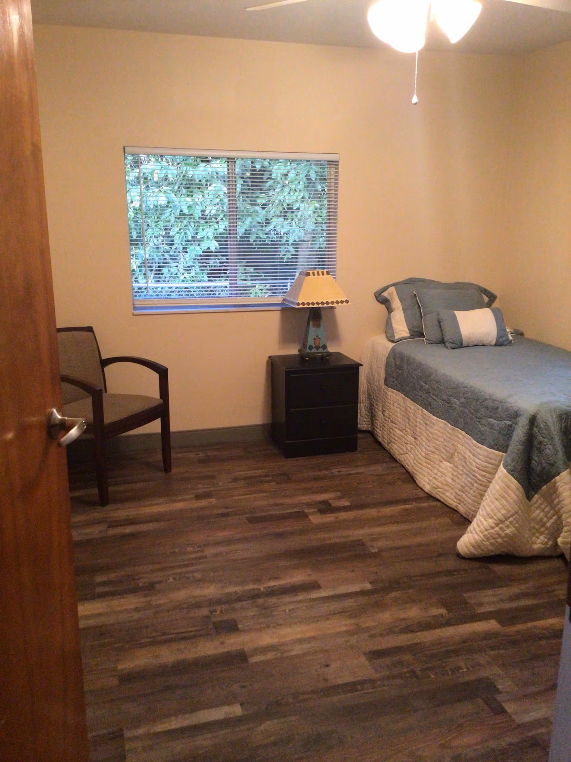 A small bedroom with a single bed covered in a blue and white quilt, a matching pillow set, a black nightstand with a decorative lamp, a wooden chair with armrests, and a window with blinds showing green foliage outside. The room has wooden flooring and beige walls.