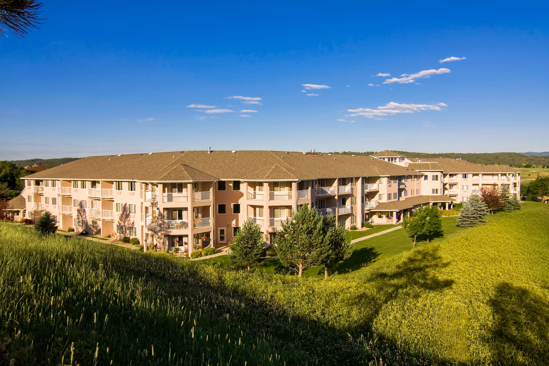 Exterior view of a large, multi-story residential building surrounded by green grass, trees, and a clear blue sky with a few clouds.