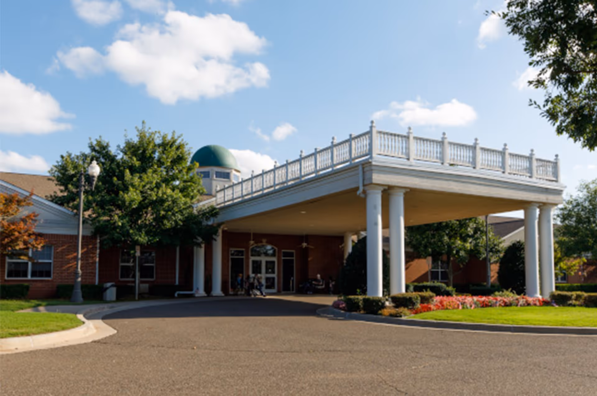 Exterior view of Arbor House of Midwest City showing the front entrance with a large covered driveway supported by white columns, surrounded by green trees, bushes, and colorful flowers under a partly cloudy blue sky.