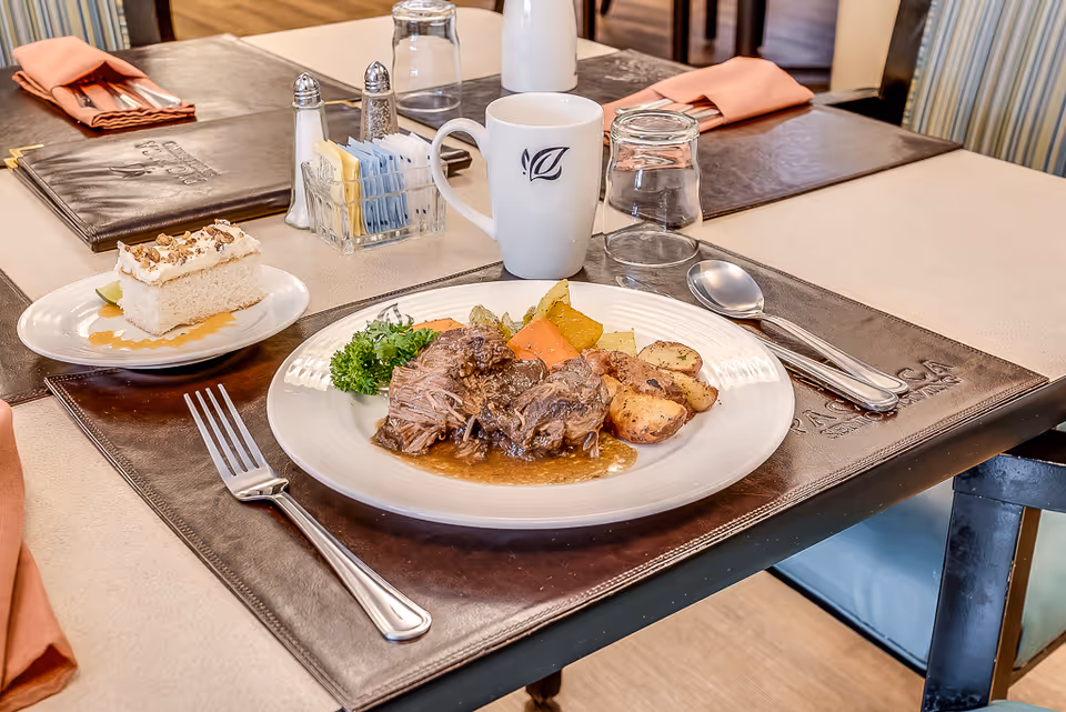A plated meal of pot roast with roasted potatoes and vegetables, a slice of cake, mug and utensils set on a dining table.