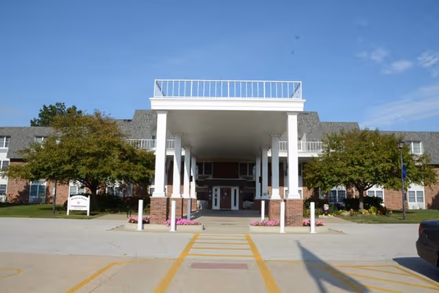 Front exterior view of Wesley Village Retirement Center showing a large covered entrance with white columns and brick bases, flanked by trees and a clear blue sky.