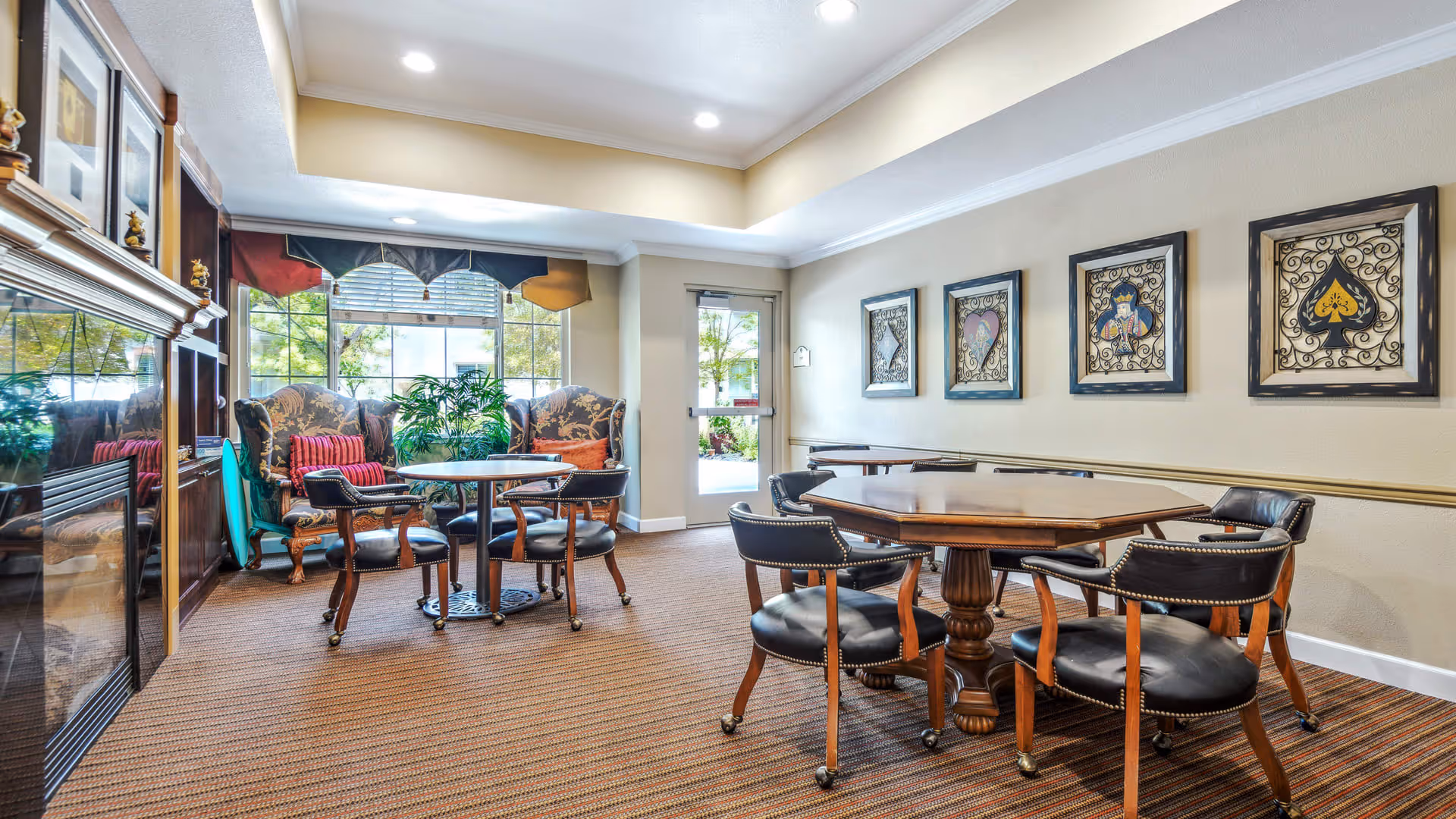 A cozy common area in a senior living facility with two wooden tables surrounded by black leather chairs on wheels. There are two upholstered armchairs with patterned fabric and red cushions near a large window with a valance. The walls are decorated with framed artwork featuring playing card suits. The room has a carpeted floor and a door leading outside.