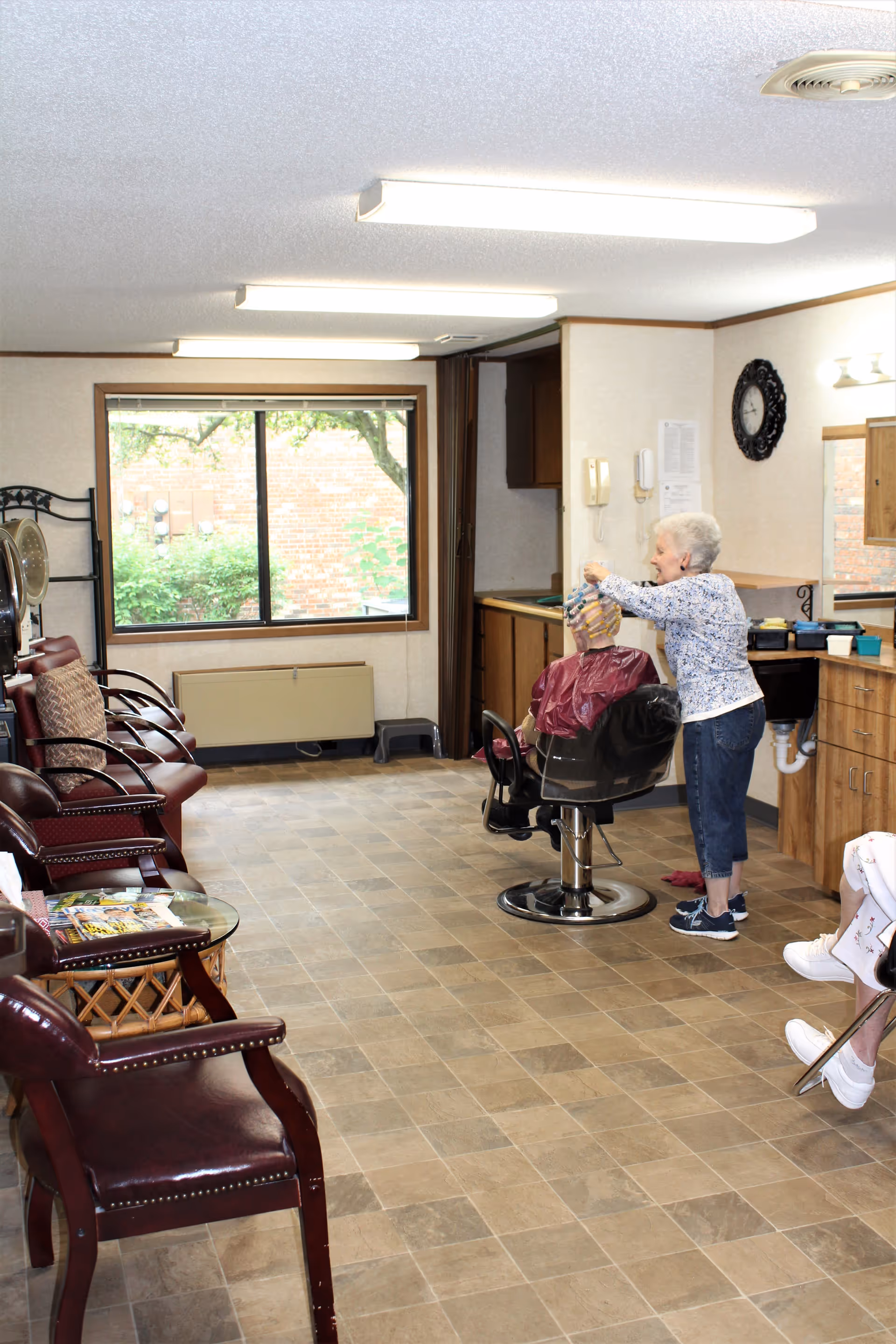 Interior of a hair salon area in a retirement facility with a woman styling another woman's hair who is seated in a salon chair. There are several empty chairs lined up along the left wall, a large window showing greenery outside, and a clock on the wall above a counter with a sink.