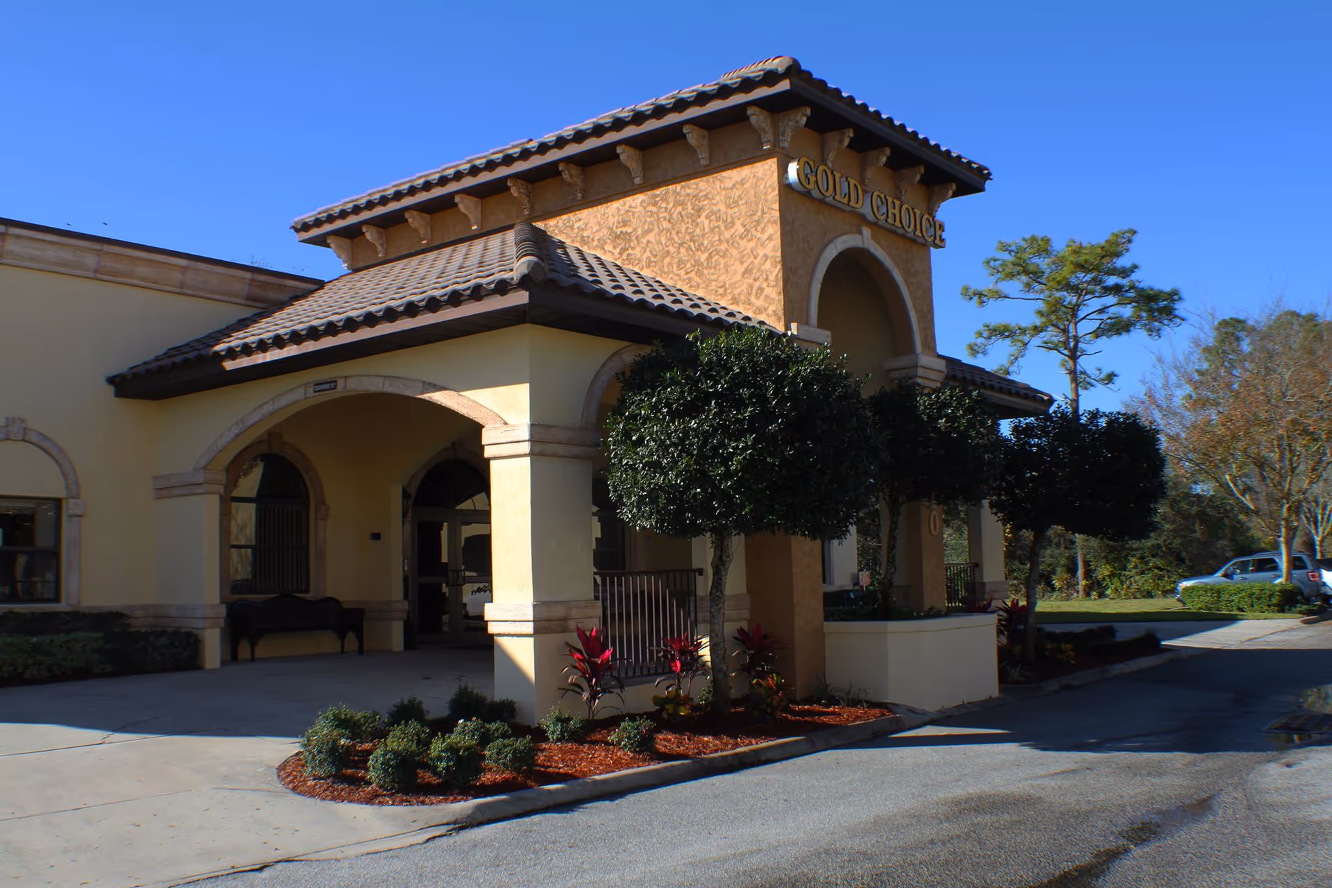 Exterior view of Gold Choice Ormond Beach Assisted Living facility showing the entrance with an arched covered driveway, manicured bushes, and trees under a clear blue sky.