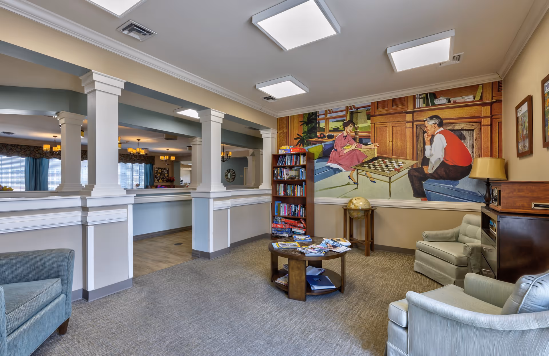 A cozy common area in a senior living facility with comfortable armchairs, a round wooden coffee table with magazines, a bookshelf filled with books and board games, and a large mural on the wall depicting a man and woman playing checkers. The room has soft lighting and an open view into an adjacent dining area with windows and chandeliers.