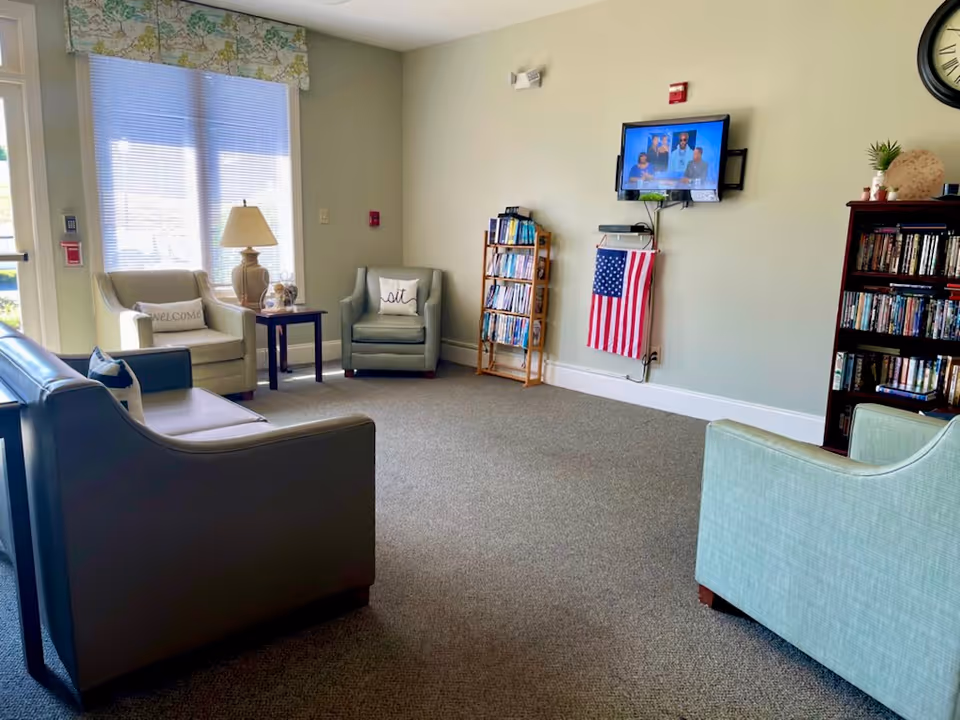 A cozy living room area with several armchairs and a sofa arranged around a carpeted floor. There is a side table with a lamp near a window with blinds and floral curtains. A wall-mounted TV is on the far wall above a small bookshelf and an American flag hanging below it. Another bookshelf filled with DVDs or books is on the right side, and a large clock and small plant are on top of it.