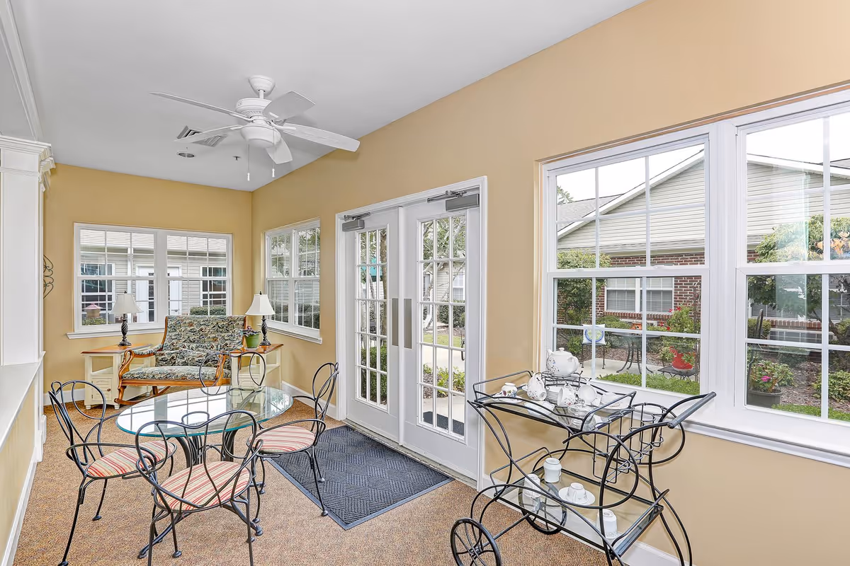 A bright sunroom with beige walls and large windows showing an outdoor garden area. The room features a glass-top round table with four wrought iron chairs with striped cushions, a floral upholstered loveseat with wooden arms, two table lamps on side tables, a ceiling fan, and a metal tea cart with a white tea set.