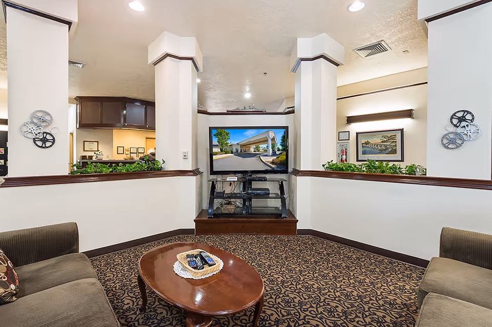 A cozy living room area in Inland Point Retirement Community featuring two brown upholstered sofas facing a flat-screen TV mounted on a stand. The room has patterned carpet flooring, white walls with dark wood trim, decorative film reels on the walls, and green plants along the half walls. The TV displays an image of the building's exterior entrance.