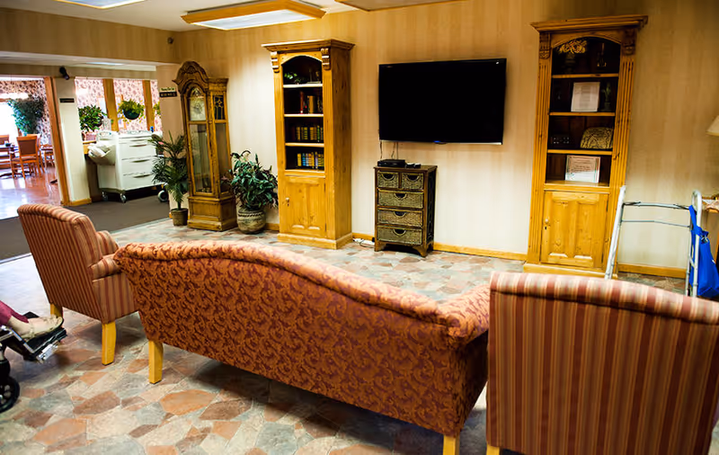 A cozy living room area in a senior living facility with a patterned sofa and two striped armchairs facing a wall-mounted flat screen TV. The room features wooden bookshelves, a small chest of drawers, a grandfather clock, and some potted plants. In the background, there is a dining area visible through an open doorway.