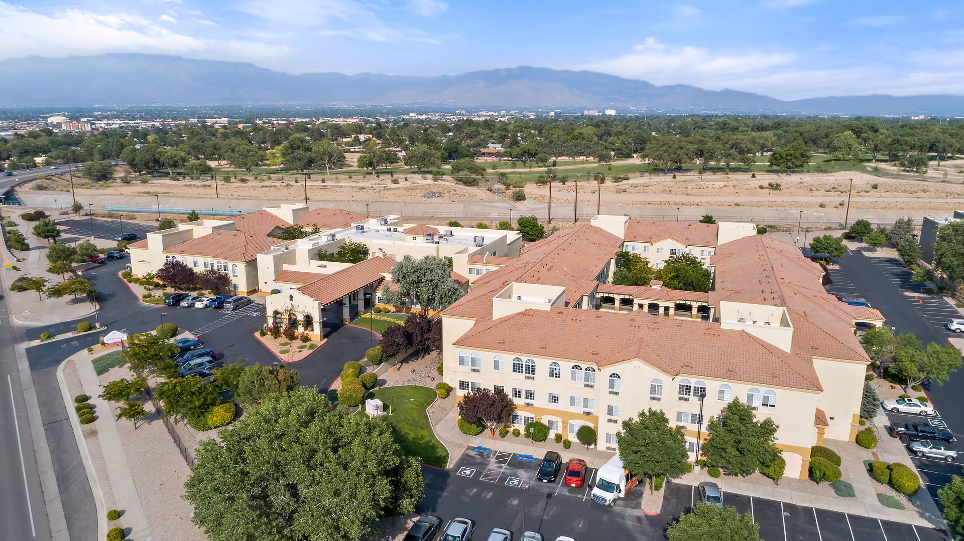 Aerial view of Morada Albuquerque senior living facility showing multiple beige buildings with red-tiled roofs, surrounded by parking lots, trees, and landscaped areas. Mountains and a clear blue sky are visible in the background.