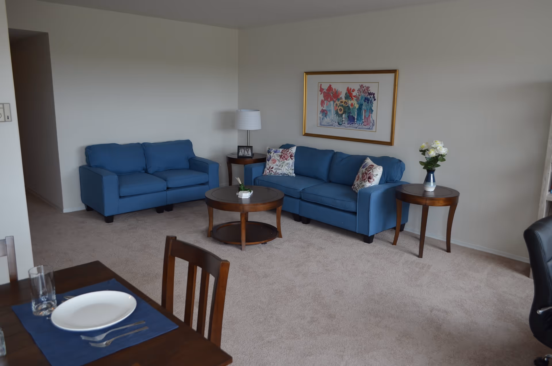 A living room area with two blue sofas, a round wooden coffee table, two wooden side tables with a lamp and a vase of flowers, a framed floral painting on the wall, and part of a dining table set with a plate, glass, and utensils.
