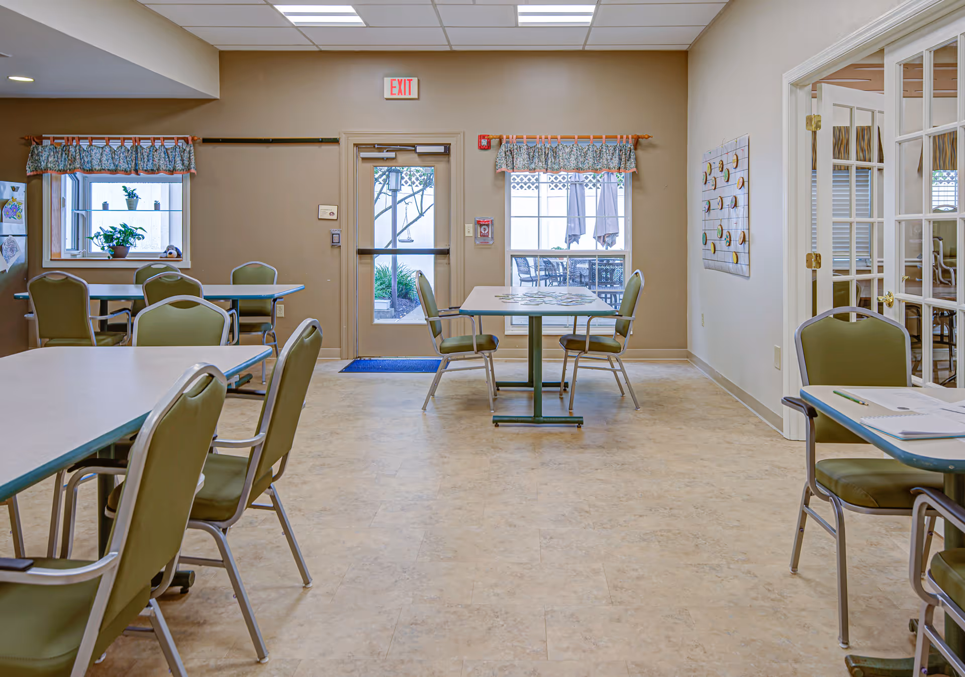 A bright room with several tables and green chairs arranged around them. There are windows with floral valances, a glass door leading outside, and a wall-mounted board with circular objects. The floor is light-colored, and the ceiling has recessed lighting.
