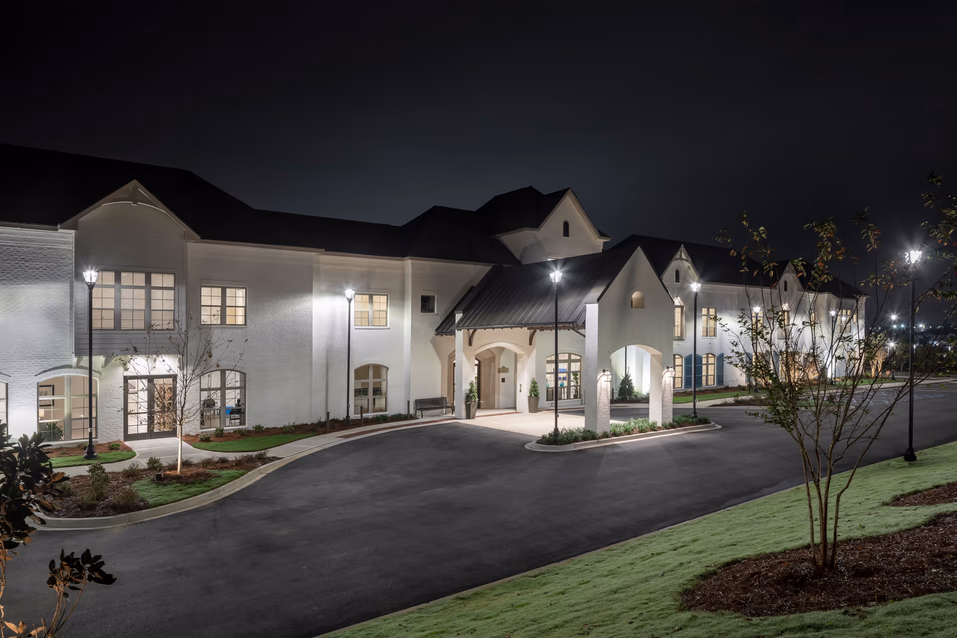 Night view of the exterior front entrance of a large, well-lit senior living facility building with white walls, multiple windows, and a covered entryway. The driveway curves in front of the entrance, and there are street lamps and landscaped greenery around the building.