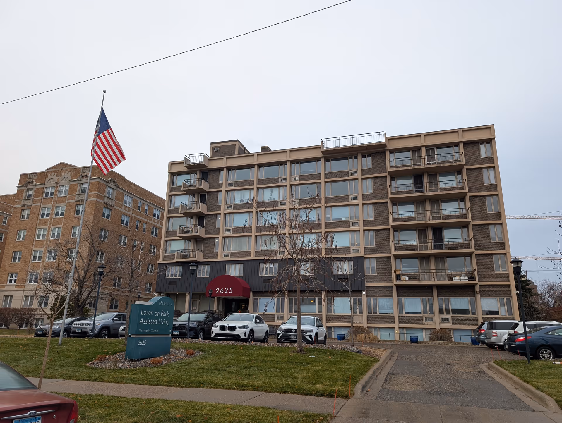 Exterior view of Loren On Park Assisted Living building with multiple floors and balconies. There is a red awning above the entrance displaying the number 2625. Several cars are parked in front of the building, and an American flag is flying on a flagpole near the entrance. A green sign in front reads 'Loren on Park Assisted Living Minneapolis Campus 2625'.