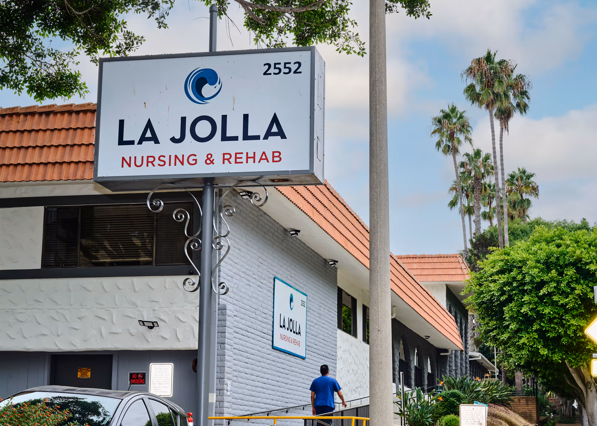 Exterior view of La Jolla Nursing and Rehabilitation Center showing a large sign with the facility name and address number 2552. A person in a blue shirt is walking near the building, which has a tiled roof and white and gray walls. Palm trees and greenery are visible in the background under a partly cloudy sky.