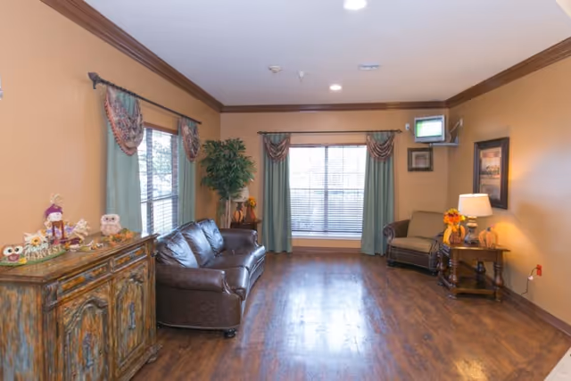 A cozy living room with wooden flooring, a brown leather sofa, a cushioned armchair, a wooden side table with a lamp and fall-themed decorations, a large window with green curtains and valances, a wooden cabinet with decorative items including owls and a scarecrow, and a small wall-mounted TV in the corner.