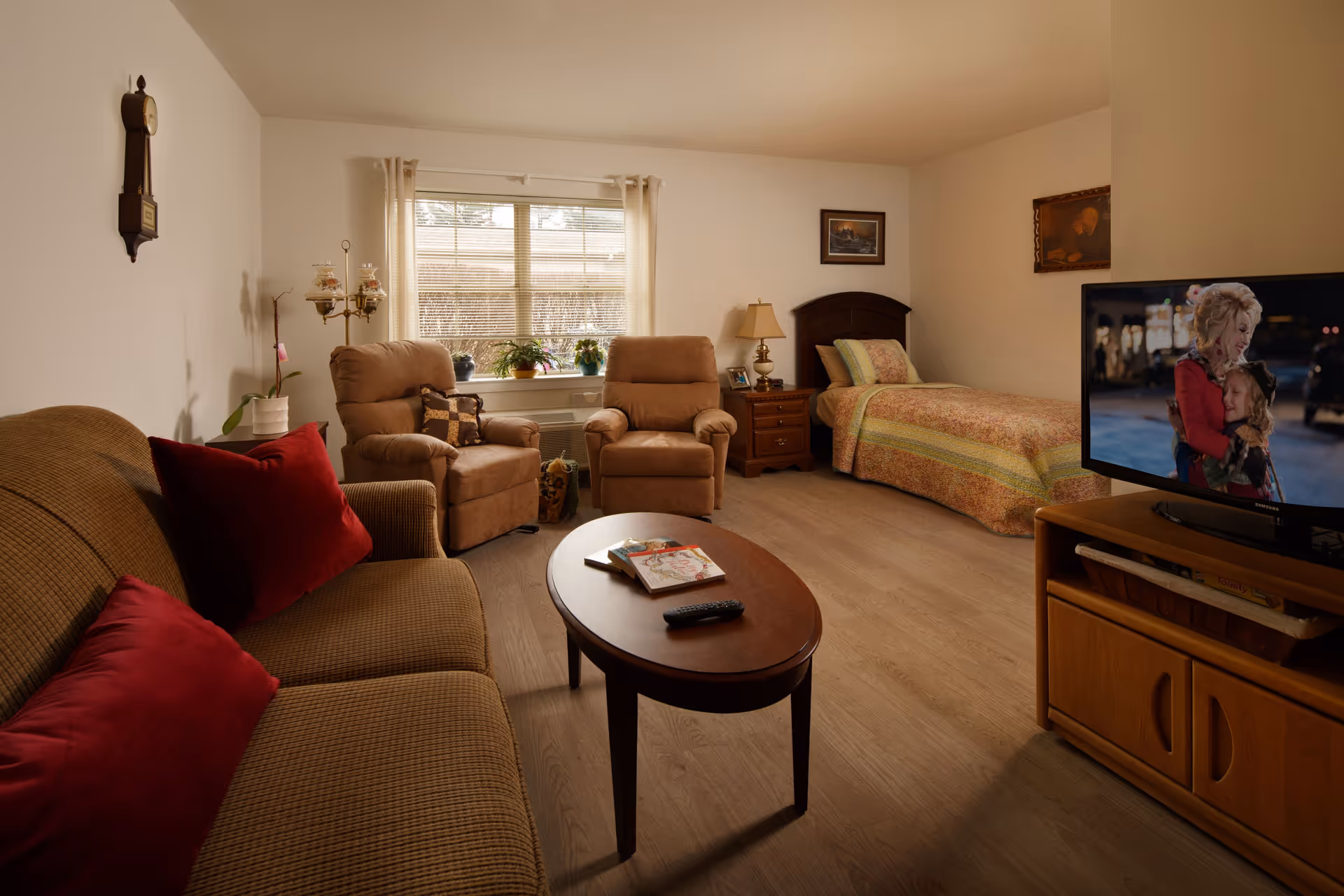 A cozy room with a brown sofa adorned with red pillows, two beige armchairs near a window with plants on the windowsill, a wooden coffee table with books and a remote control, a single bed with a patterned bedspread, a nightstand with a lamp, and a television showing a scene of a woman hugging a child.