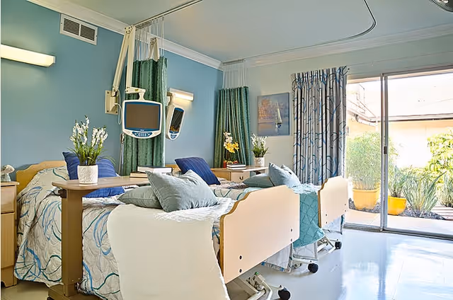 Two adjustable hospital beds in a bright, decorated patient room with monitors, bedside tables, green curtains, and a sliding glass door to a patio.