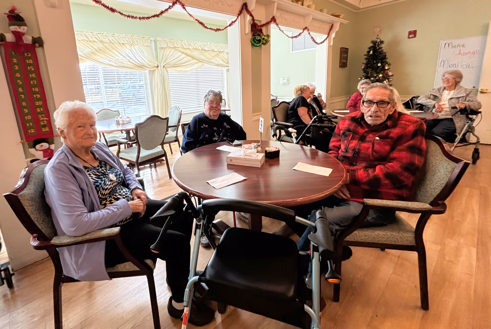 Elderly residents seated around a table in a holiday-decorated common dining area of a senior living facility.