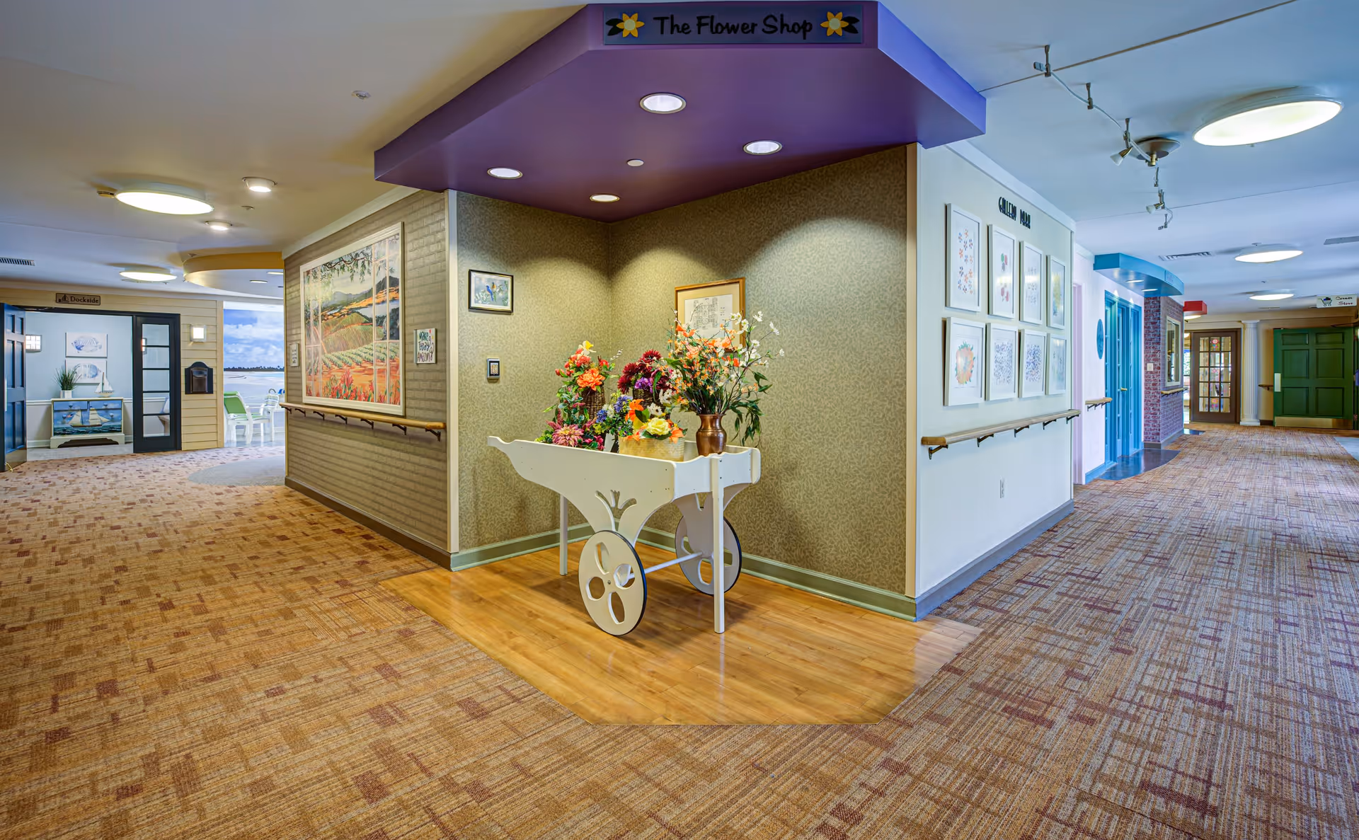 Interior hallway of a senior living facility with carpeted floors and handrails along the walls. A white flower cart labeled 'The Flower Shop' is positioned in a corner, displaying colorful artificial flowers. The hallway features framed artwork on the walls and leads to various rooms with different colored doors. There is a seating area visible in the distance near large windows.