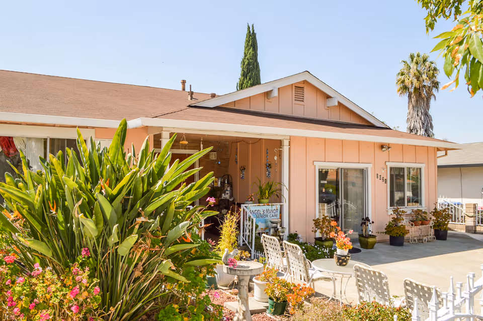 Exterior view of a single-story pink senior care facility with a porch, potted plants, outdoor chairs, and a garden with green foliage and colorful flowers under a clear blue sky.