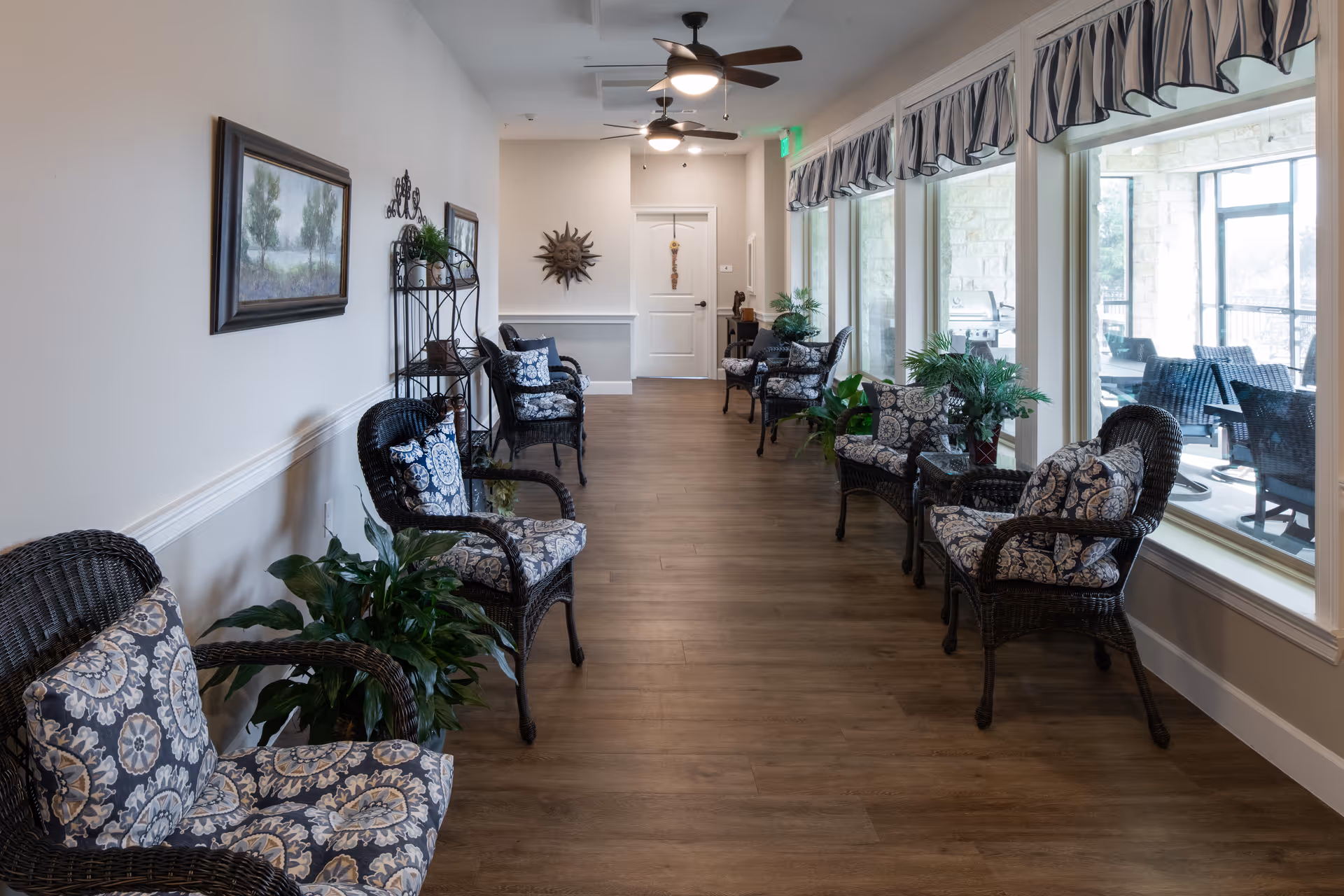 A long interior sitting area with wicker chairs and patterned cushions lined along a hallway next to large windows and potted plants.