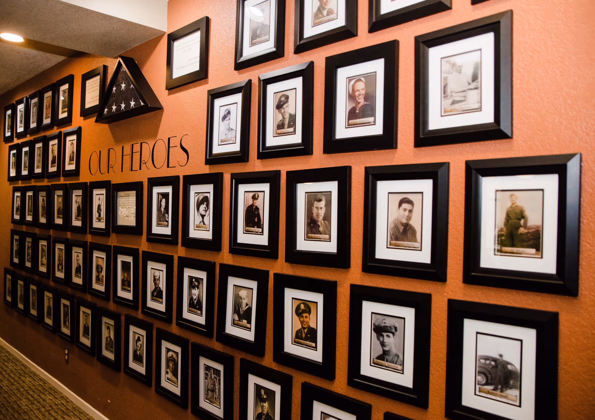 A hallway wall painted in orange with multiple framed black-and-white and sepia-toned photographs of military personnel arranged in a grid. Above the photos, the words 'OUR HEROES' are written on the wall, and a folded American flag is displayed in a triangular case.