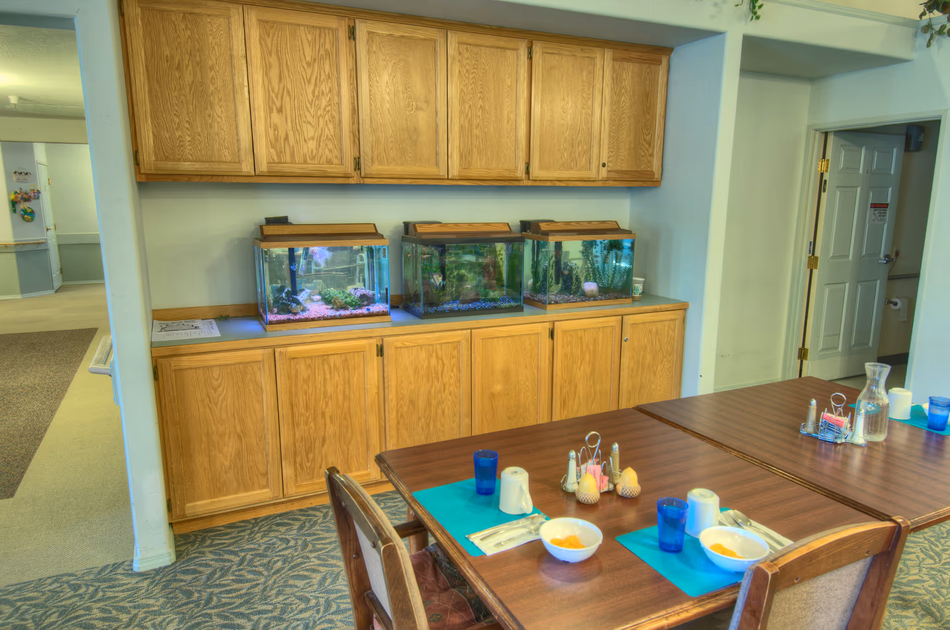 Dining area with wooden tables set with bowls, cups, and utensils on blue placemats. Behind the tables, there is a wooden cabinet with three fish tanks on top. The room has light-colored walls and carpeted flooring with a patterned design. A doorway is visible leading to another room.