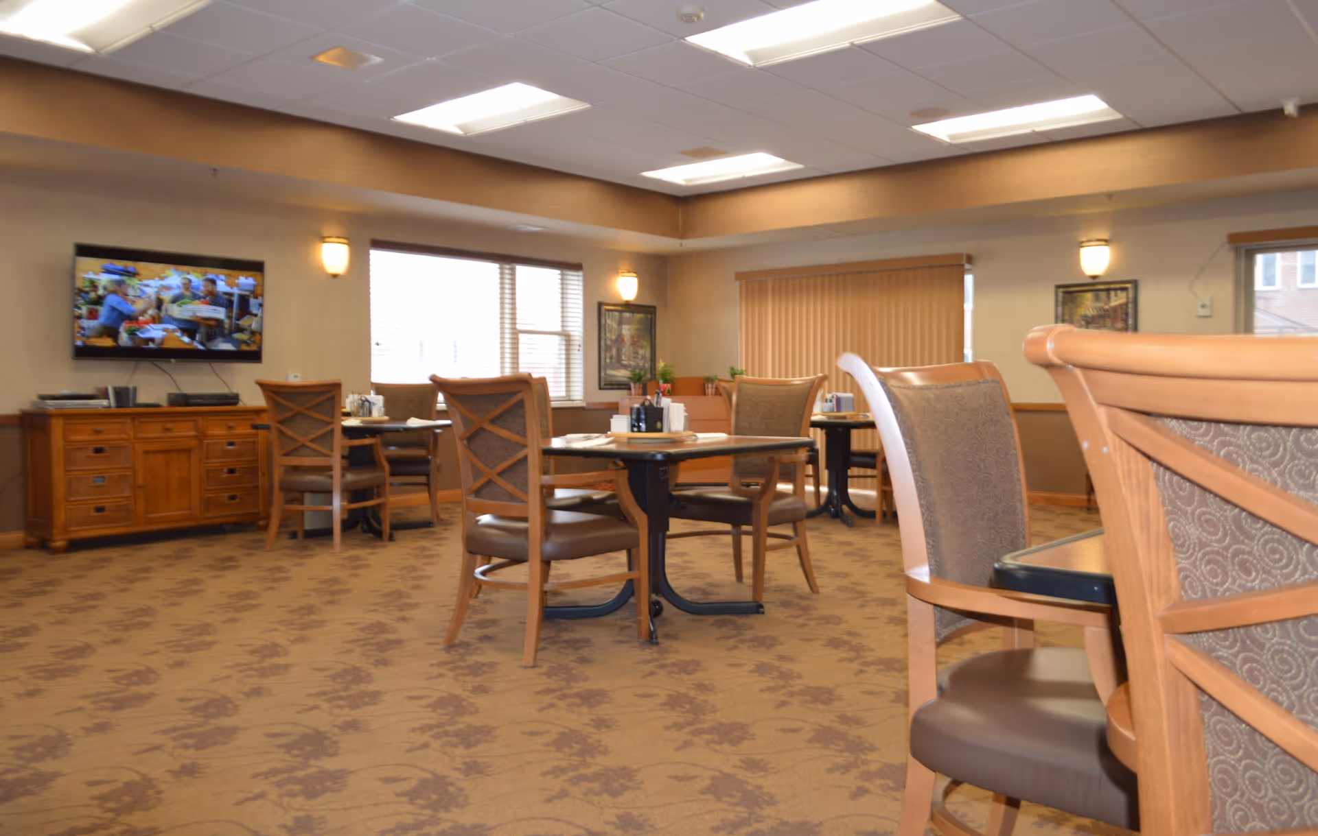 Spacious assisted living dining room with wooden tables and chairs, a TV on a sideboard, and large windows.