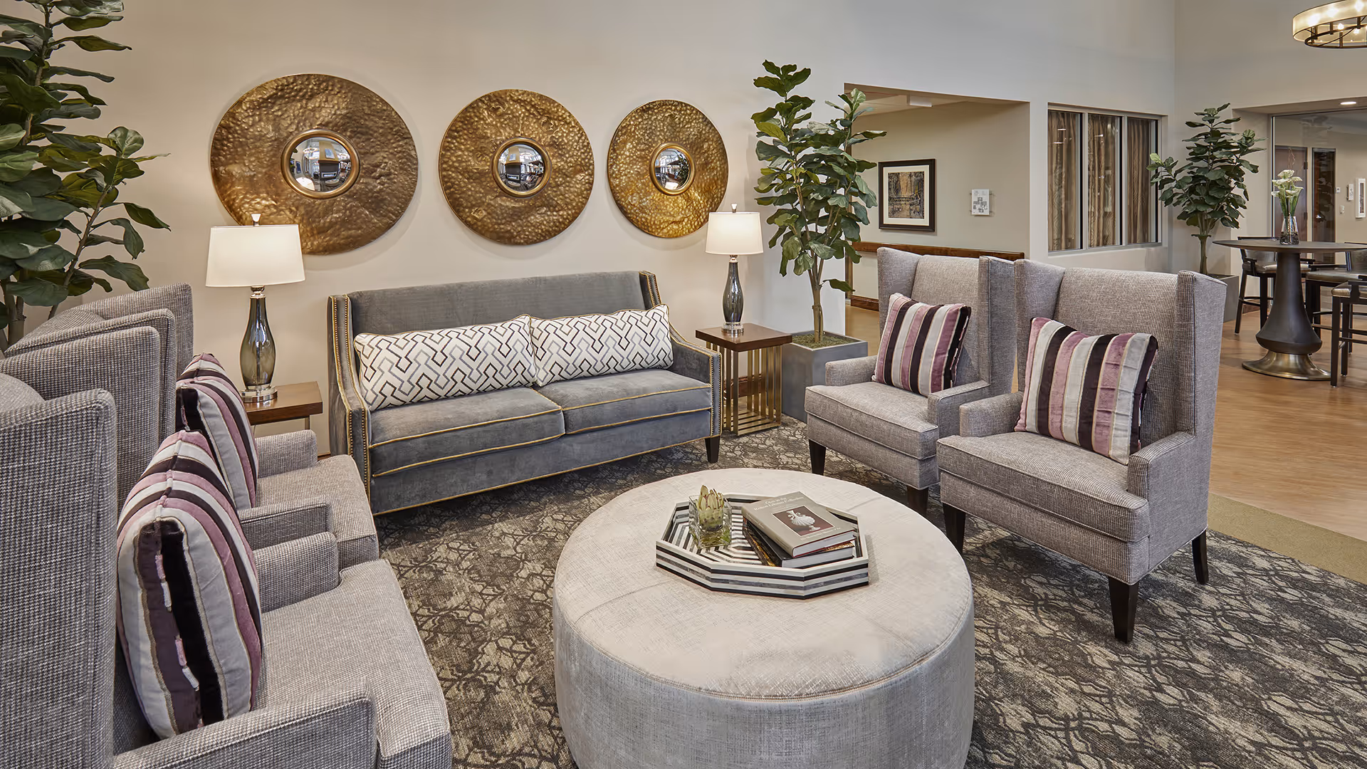 A cozy and stylish living room area in a senior living facility featuring a gray sofa with patterned cushions, two gray armchairs with striped pillows, a large round ottoman with books and a decorative tray on top, two side tables with lamps, three large round metallic wall decorations, and several potted plants. The room has a patterned carpet and wooden flooring in the background with additional seating and decor.