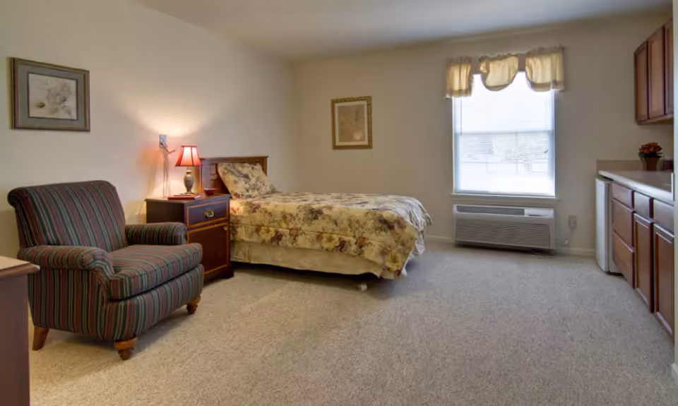 A senior living facility room with a single bed covered in floral bedding, a striped armchair, a wooden nightstand with a lamp, framed artwork on the walls, a window with a valance, and a kitchenette area with cabinets and a mini refrigerator.