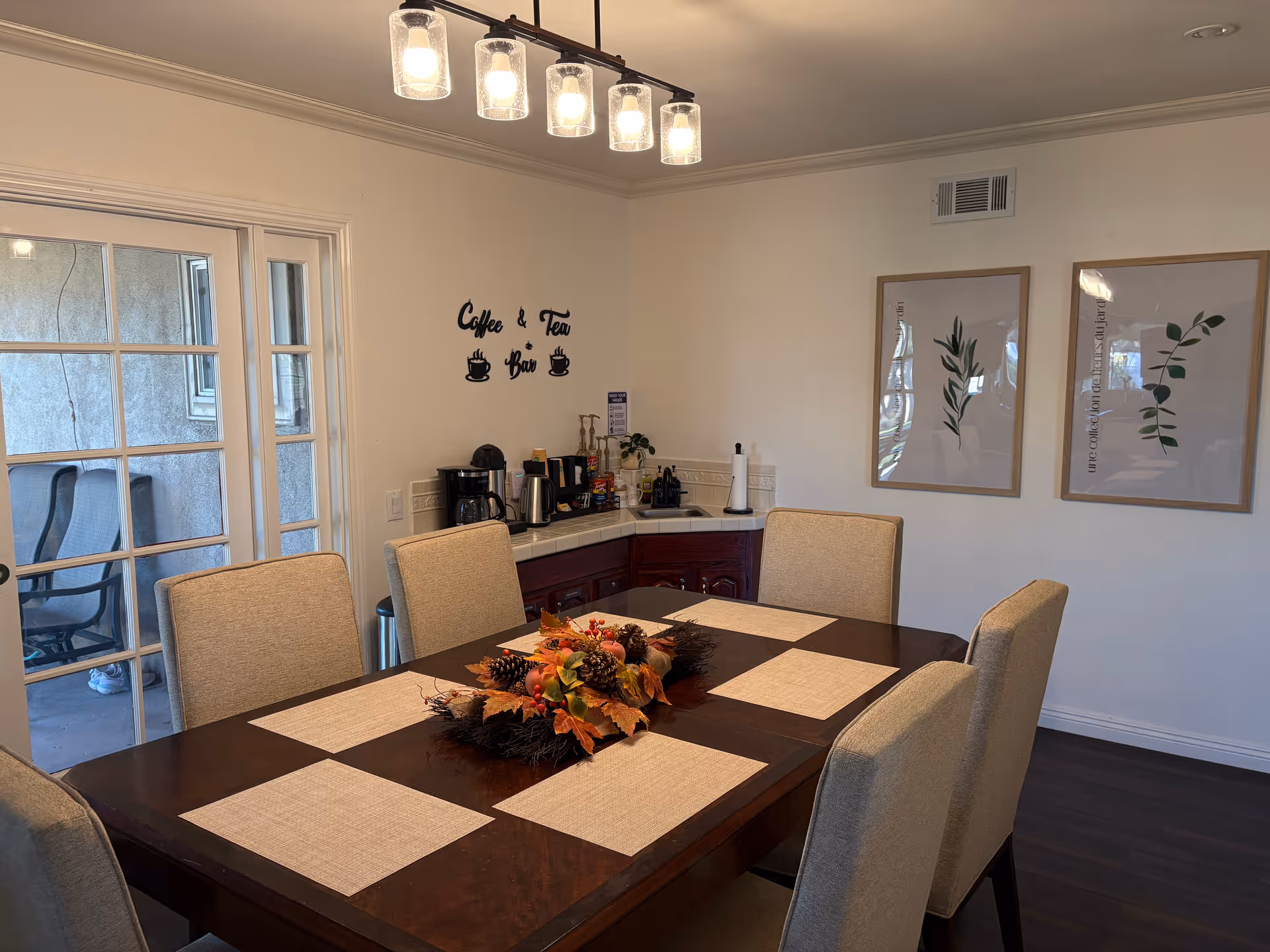 Dining room with a large wooden table set with placemats and a seasonal centerpiece, upholstered chairs, a coffee and tea station against the wall, and framed botanical prints.