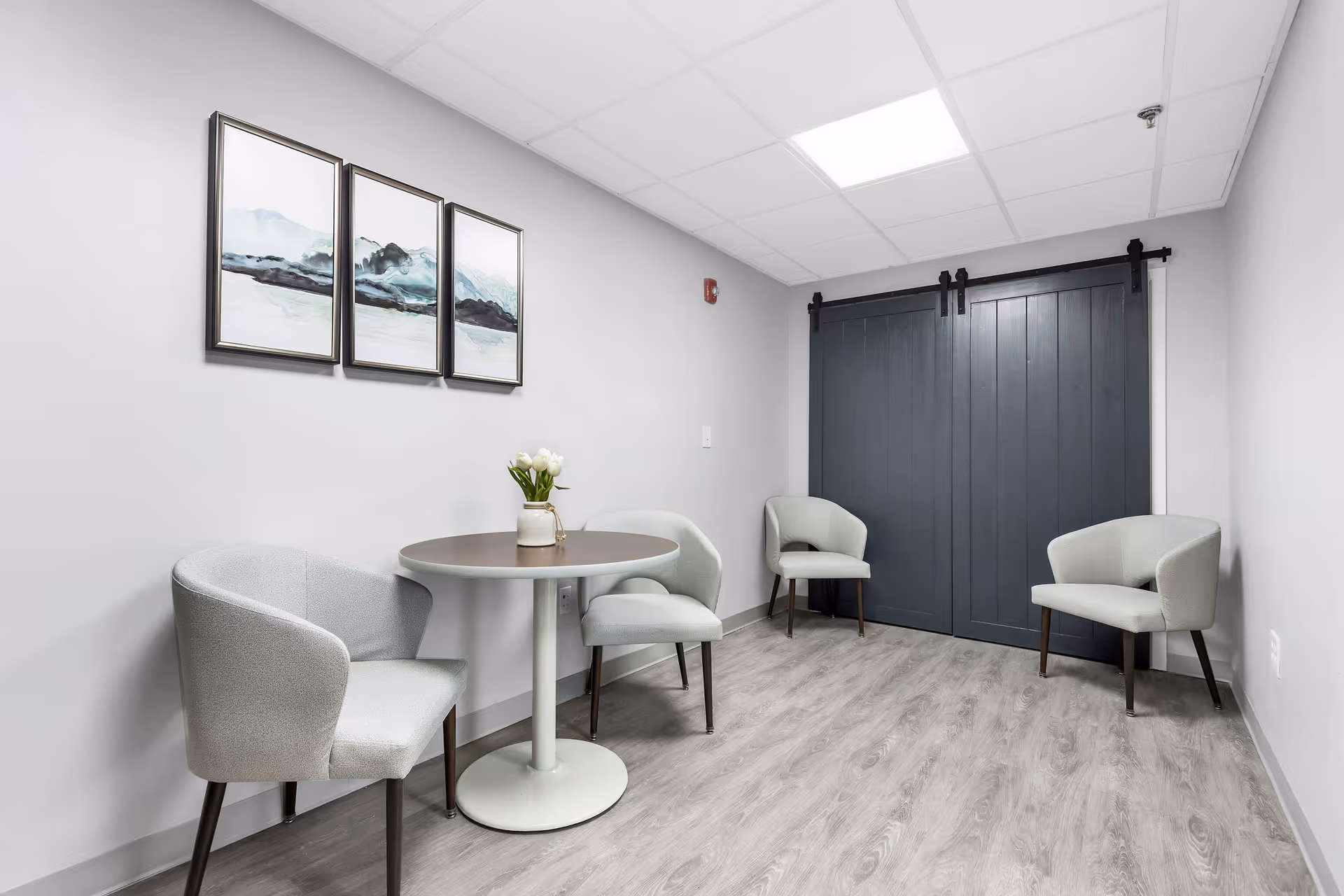 Small seating area with a round table, four gray upholstered chairs, framed wall art, and dark sliding barn doors.