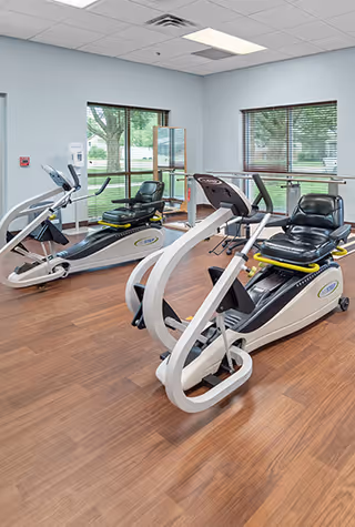 A bright exercise room with two recumbent stationary bikes positioned on a wood floor. The room has large windows with blinds, allowing natural light to enter. The walls are painted light blue, and there is a hand sanitizer dispenser mounted on the wall near the window.