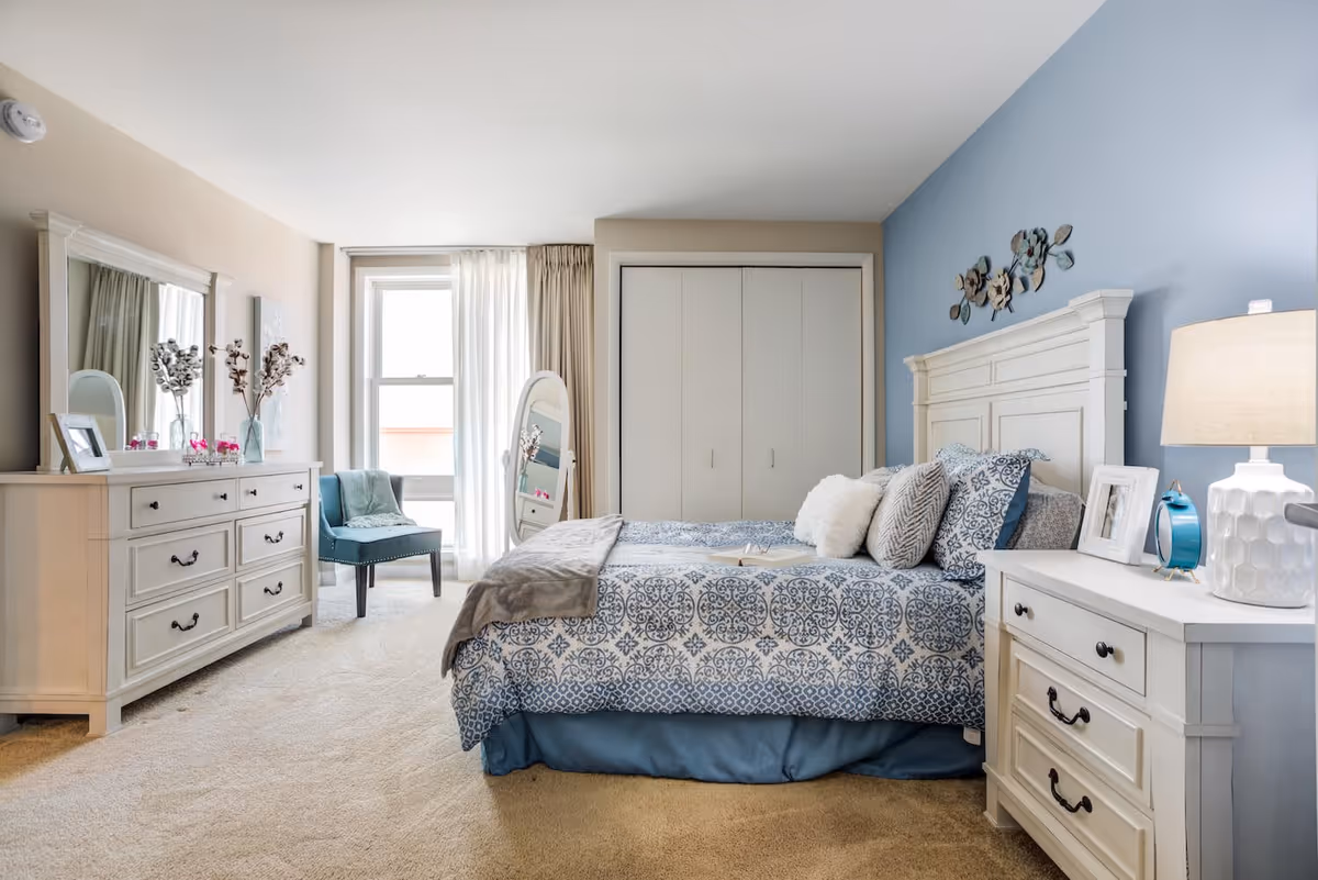 A bright and cozy bedroom featuring a large bed with blue and white patterned bedding and multiple pillows. The room has a white dresser with a mirror, a small white nightstand with a lamp and decorative items, a blue accent wall with floral wall art, a blue upholstered chair near a window with sheer curtains, and a full-length mirror leaning against the wall.
