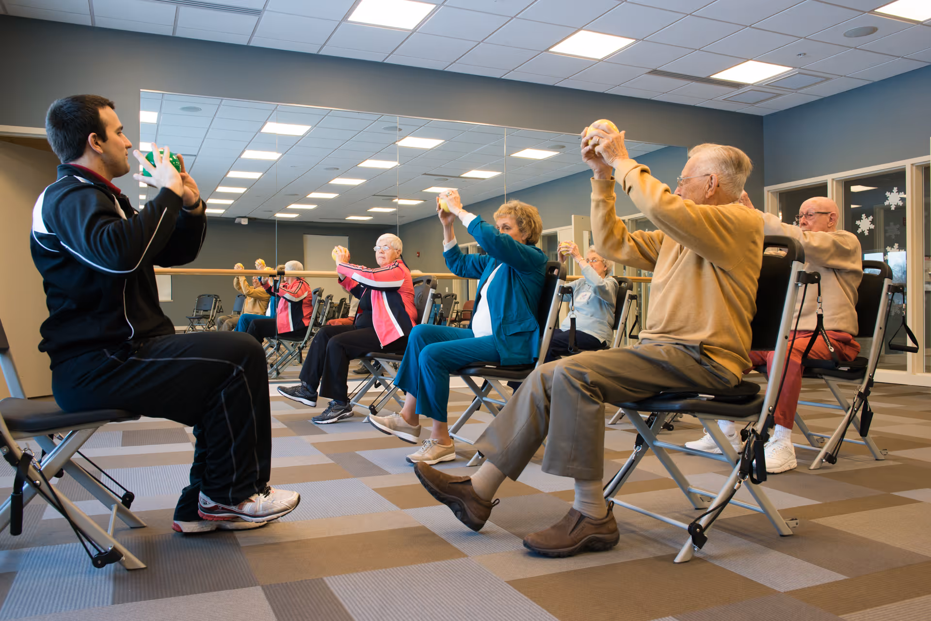 A group of elderly individuals seated on chairs in a fitness room participating in a seated exercise class led by an instructor. They are holding small exercise balls and stretching their arms forward. The room has a large mirror on one wall and a checkered carpet floor.
