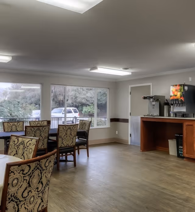 A dining area with several tables and patterned chairs, large windows showing parked cars and greenery outside, and a beverage station with a coffee machine and a soda dispenser on a wooden counter.