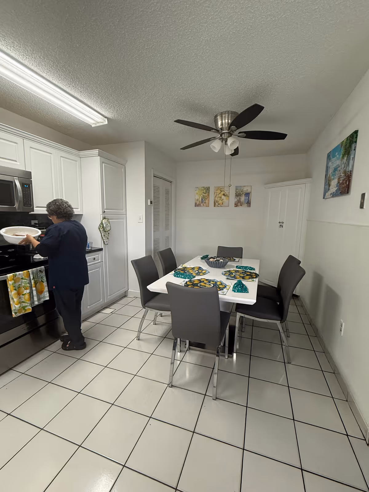 A woman is standing in a kitchen area near a stove, holding a bowl. The kitchen has white cabinets, a microwave, and a black stove. There is a dining table with six gray chairs around it, set with colorful placemats and a bowl in the center. The room has white tiled flooring, a ceiling fan with lights, and artwork on the walls.