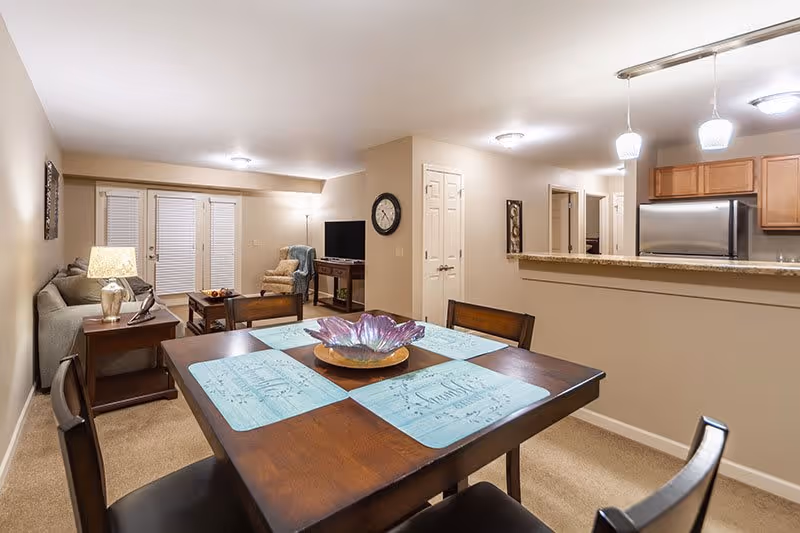 Interior view of a senior living facility showing a dining area with a wooden table set with four blue placemats and a decorative bowl in the center. Beyond the dining area is a living room with a sofa, side table with a lamp, an armchair, a TV on a stand, and a clock on the wall. The kitchen area with wooden cabinets and stainless steel refrigerator is visible to the right, separated by a counter with pendant lights hanging above.