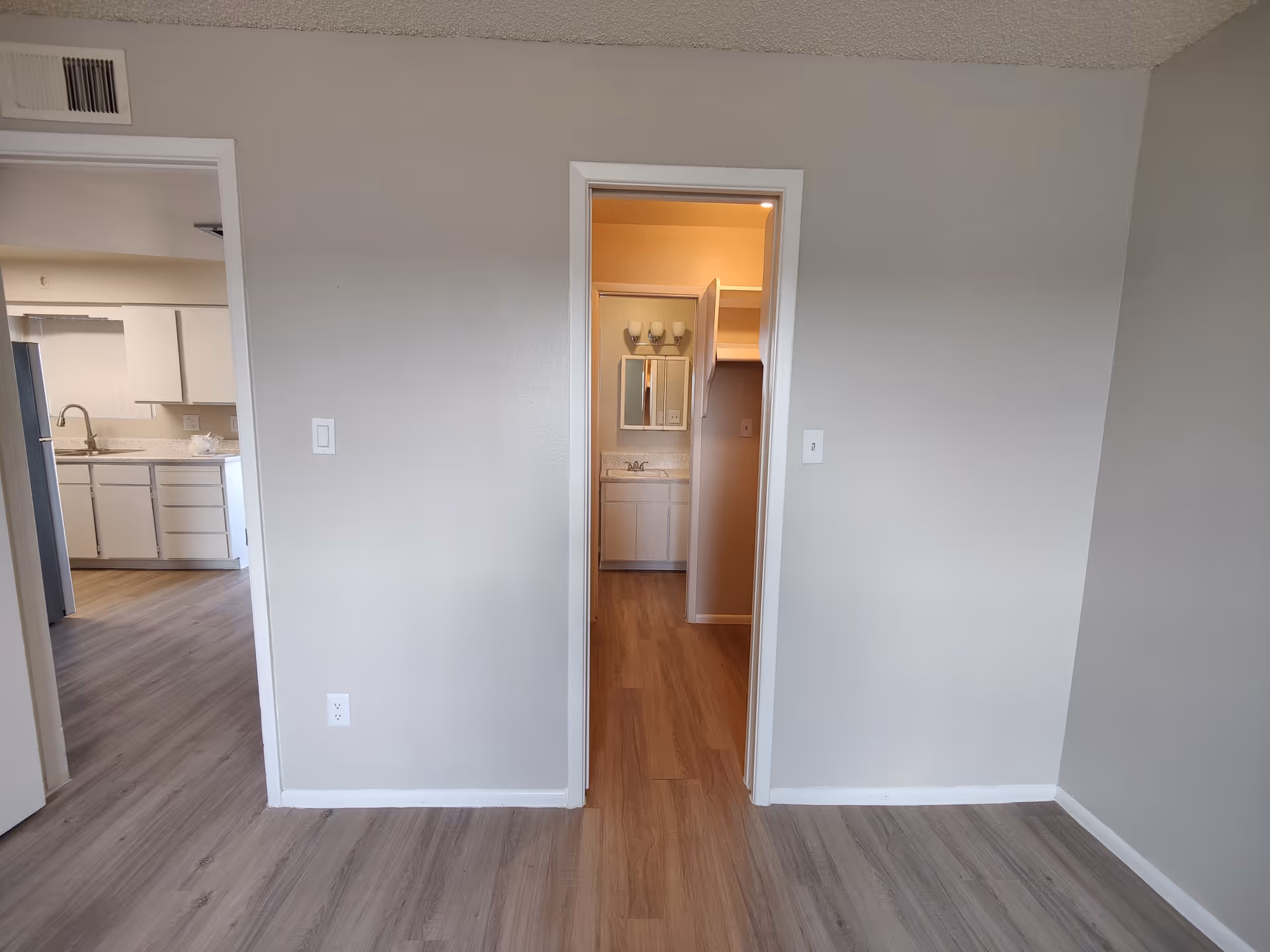 View of an interior space showing two doorways. The left doorway leads to a kitchen area with white cabinets, a countertop, and a sink. The right doorway leads to a bathroom with a vanity, mirror, and light fixtures above the mirror. The floors are wood-style laminate and the walls are painted light gray.