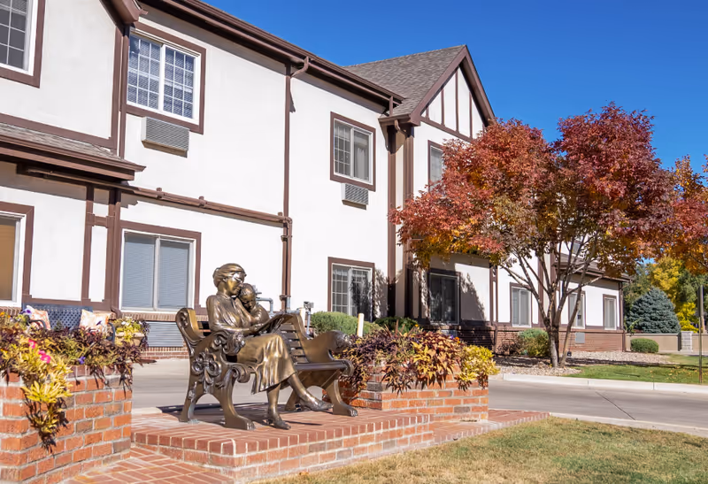 Outdoor view of a senior living facility building with white walls and brown trim under a clear blue sky. In the foreground, there is a bronze statue of a woman reading to a child seated on a bench, surrounded by brick planters with colorful plants. A tree with red and orange autumn leaves is visible near the building.