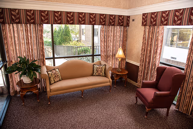 A cozy sitting area with a beige upholstered sofa adorned with two patterned cushions, a burgundy armchair, two wooden side tables, one holding a potted plant and the other a lamp, all set against large windows with patterned curtains and valances, allowing natural light to brighten the room.